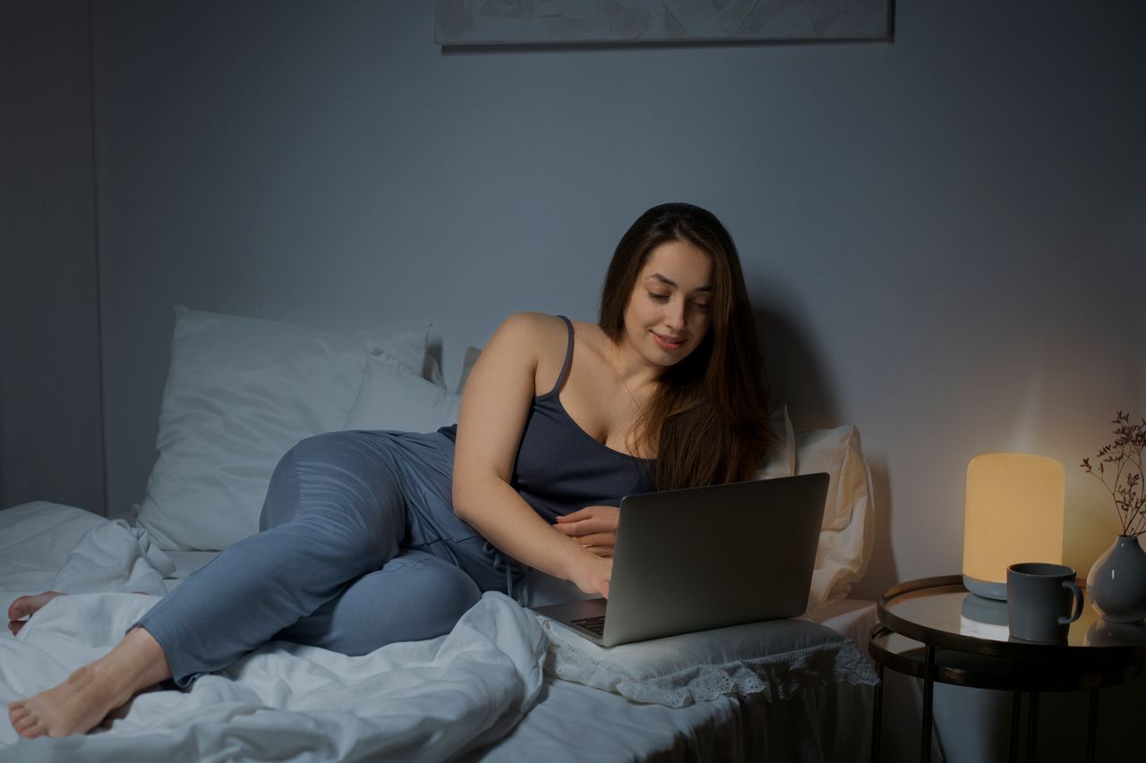 A woman lounges on her bed and smiles at her laptop