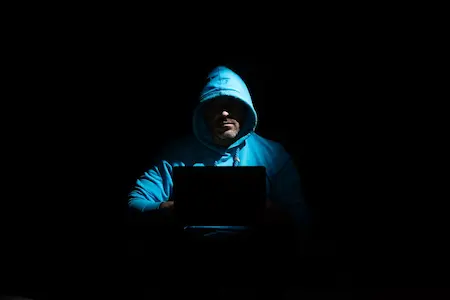 A suspicious man wearing a hoodie using a laptop in the dark