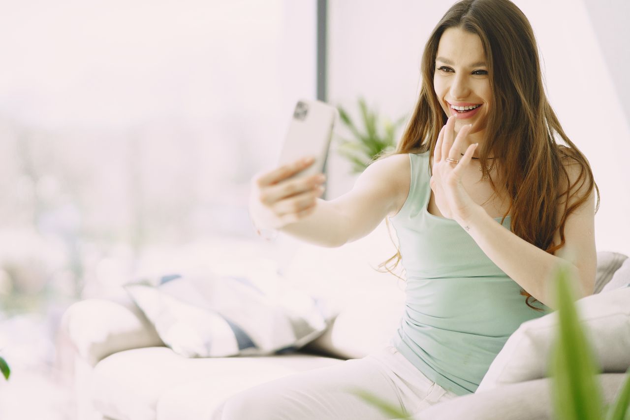 A woman sitting on her couch while waving and smiling at her cell phone